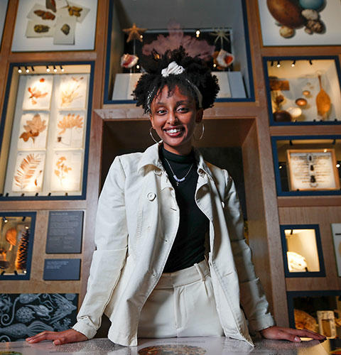 A female student with a natural afro hairstyle wears a white jacket and stands in a museum-like exhibit of natural specimens, smiling at the camera.