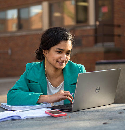 A female student with dark hair, wearing a turquoise jacket, sits at an outdoor table studying with a laptop, books, and a smartphone, smiling slightly.