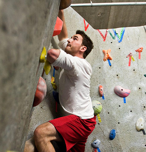 A male student with short hair wearing a white shirt and red shorts is climbing an indoor rock wall, gripping colorful handholds.