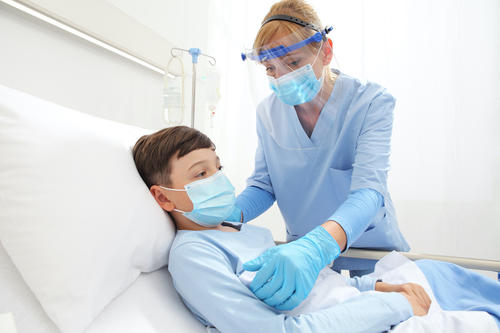 Nursing wearing face shield helping young boy wearing mask in hospital bed