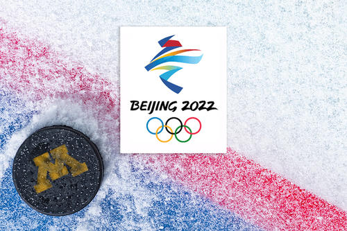 A hockey puck with a gold M on it on a sheet of ice with the Beijing Olympics logo