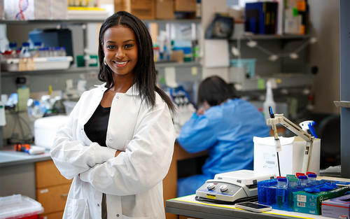 Abygail Andebrhan crossing arms while smiling in a white lab coat standing in a lab