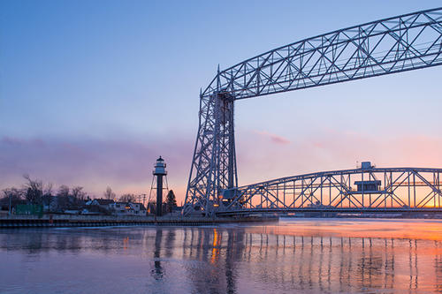 Photo of sunset reflected in Lake Superior. Visible across the water is a lighthouse and the Aerial Lift Bridge—landmarks of Duluth, Minnesota.