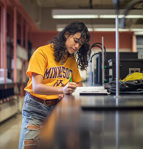 Female student wearing gold UMN shirt writes in notebook in lab setting