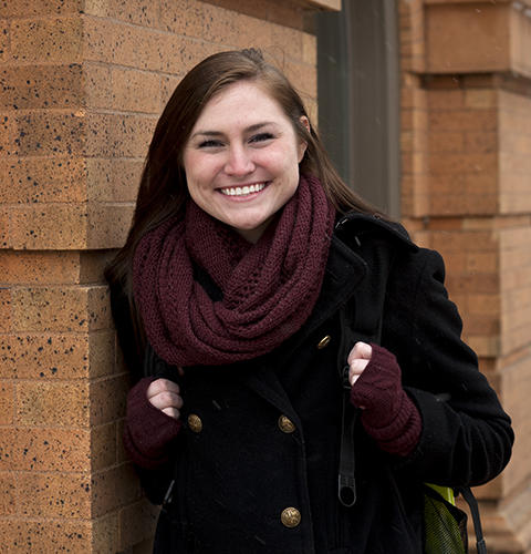 A female student smiles while she leans against a brick campus building wearing warm clothing
