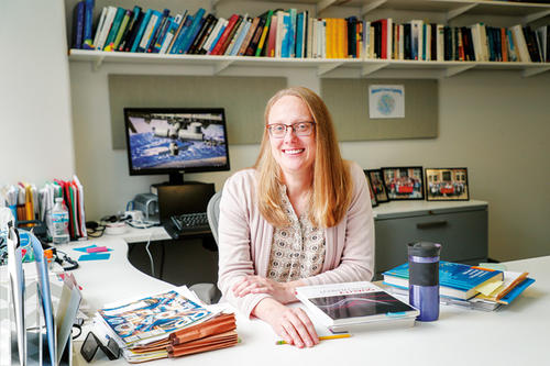   Lisa Miller, blonde, long hair, glasses, high forehead, wears a light-colored sweater and sits at a desk with books on it.