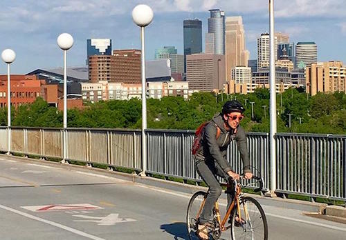 A cyclist bikes across the Washington Avenue pedestrian bridge on the University of Minnesota campus. The Minneapolis skyline is in the background.