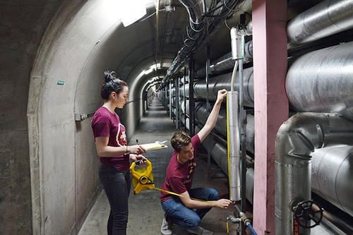 A female and male student check out a wall of large silvery pipes.