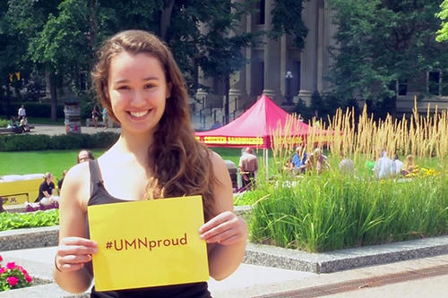 A girl standing on Northrop Plaza holding a sign that says &quot;#UMNProud&quot; 