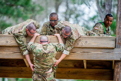 A group of soldiers, in camouflage, help another soldier over a wall in a bootcamp test.