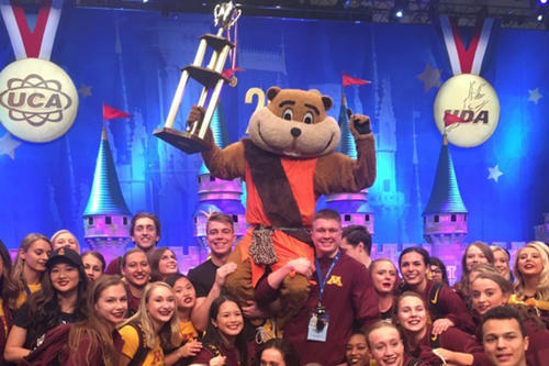 A jubilant Goldy Gopher holding a championships trophy and surrounded by U of M cheer and dance team members