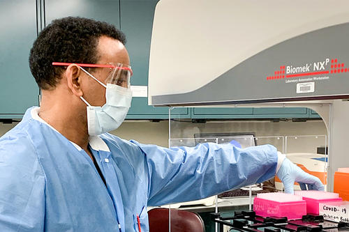 A man in short curly dark hair, in white mask and blue lab coat, reaches into a workstation to grab a container.