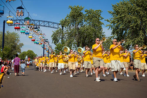 A marching band wearing gold T-shirts walks while playing at the Minnesota State Fair with the Skyride operating overhead.