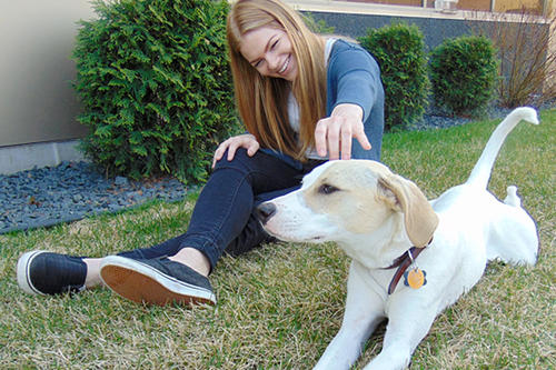 Aimee Krebsbach sits on grass reaching to pet mostly white short-haired dog.