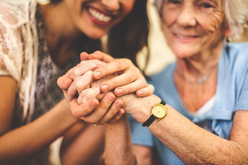 An older woman and a presumed caregiver clasp hands and smile.