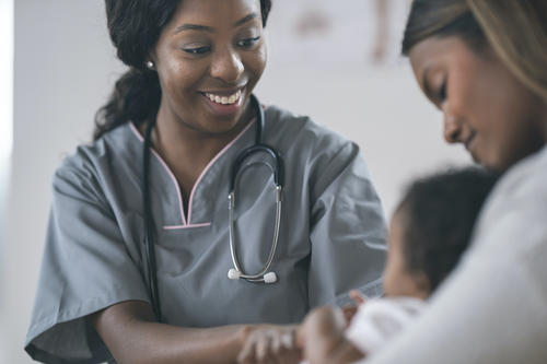 Black female physician smiling at Black newborn baby in his mom&#039;s lap