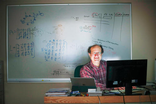 Daniel Boley, glasses, plaid shirt, balding, sits facing a computer with a blackboard behind him.