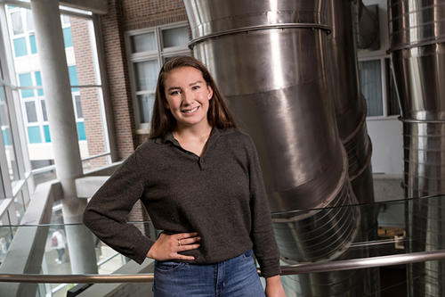 Dannyelle Donahue, dark hair, poses in front of large silver pipes in the mechanical engineering building.