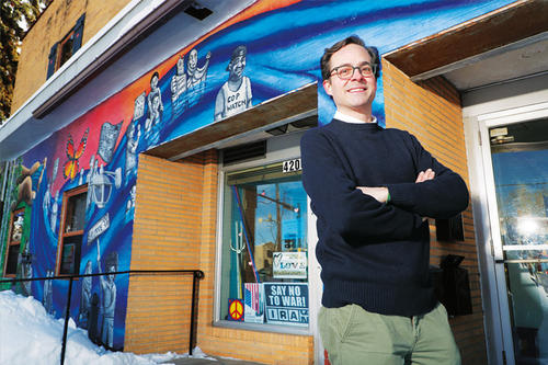 Eric Daigre, dark hair, high forehead, glasses, stands with folded arms in front of a storefront with progressive signs.