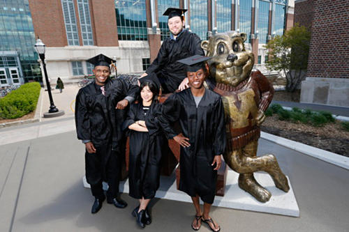 Gopher student-athletes in caps and gowns stand in front of the Goldy statue outside of the student union.