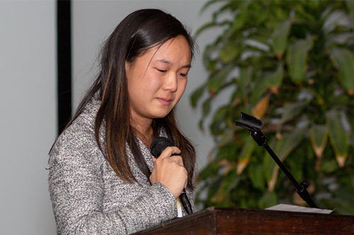 Jenny Zhang stands at a lectern, holding a mic, looking down and serious.