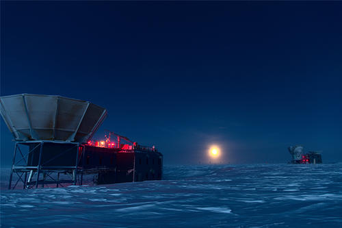 Lights on two telescopes frame a blurry moon caught between a blue expanse of snow and a dark sky. 