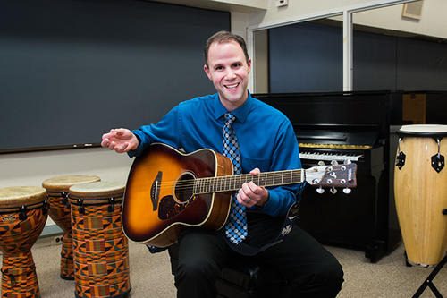 Michael J. Silverman, sitting next to timpani, smiles and holds a guitar.