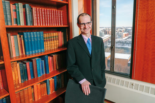 Michael Lower, in glasses and suit, stands next to a bookcase and an upper story window. 