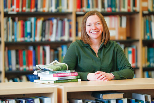 Rebecca Dean Swenson  sits at a desk before a backdrop of bookshelves.