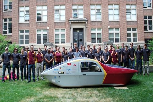 The Eos II solar car and the large team of students in front of a brick building.