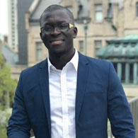 Modou Jaw wearing white shirt and blue suit jacket and glasses with gold accent, with buildings visible in background
