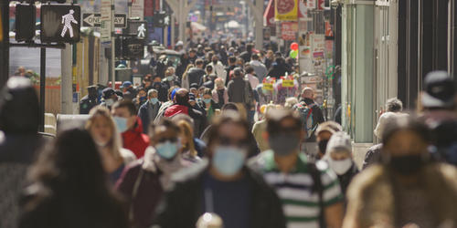 Mostly masked people, faces slightly out of focus, walk along a crowded sidewalk in New York City