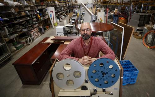 Todd Tanner, Zero Waste Program manager posing in the Reuse warehouse with and old film reel-to-reel in foreground and desks and warehouse in background