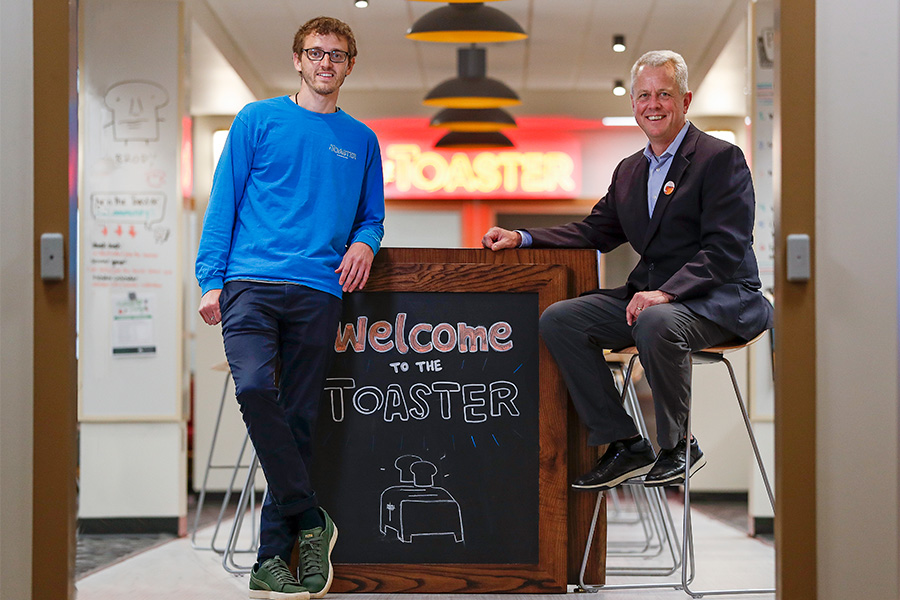 Two men posing on camera next to welcome to the toaster sign