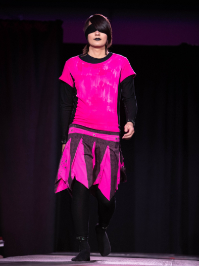 Model on runway walking toward camera wearing bright pink top with black skirt with bright pink details